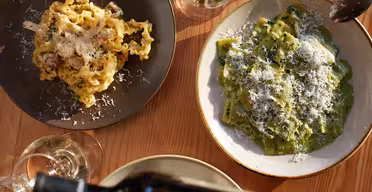 To tallerkener med pasta står på et trebord. En tallerken har båndformet pasta med en kremet saus, den andre har grønn pasta toppet med ost. Glass med vin er ved siden av dem, og en hånd heller vin fra en flaske.