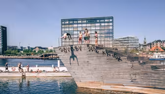 Folk dykker og sitter på en trerampe ved vannet, med moderne bygninger i bakgrunnen. Den solfylte dagen viser en livlig, rekreasjonsscene ved vannkanten.