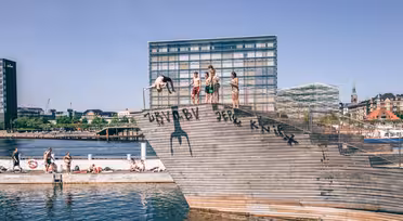 Folk dykker og sitter på en trerampe ved vannet, med moderne bygninger i bakgrunnen. Den solfylte dagen viser en livlig, rekreasjonsscene ved vannkanten.