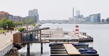 En rolig scene ved vannkanten har en liten brygge med båter bundet opp, en moderne bygning med skråtak og en bred sti med folk som går. I det fjerne er industribygg og moderne arkitektur synlig under en lett overskyet himmel.