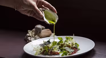 En person heller grønn saus fra et østersskall på en salat på en hvit tallerken. Salaten inkluderer bladgrønt, spiselige blomster og tynne skiver rødt kjøtt, kunstferdig arrangert. Skjell og steiner er i bakgrunnen.
