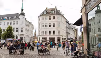 Et travelt europeisk torg med folk som går og sitter på kafeer. Historiske hvite bygninger omgir brosteinsgater. Sykler er parkert i nærheten. Et grønt spir er synlig i bakgrunnen under en overskyet himmel.