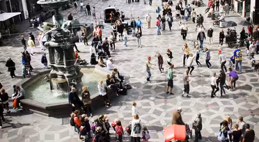 Et livlig bytorg med folk som går og samles rundt en fontene. Det torgmønstrede fortauet er omgitt av historiske bygninger. Folk blir sett på tur, tar bilder og sitter. Lyst og yrende urbant miljø.
