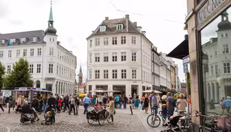 Et livlig europeisk gatebilde med folk som går og sykler på brostein. Bygningene rundt er klassiske og utsmykkede, med en fremtredende bygning med et spir. Kaféer og butikker ligger langs gaten, og flere trær gir grønt.