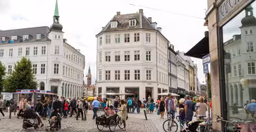 Et livlig europeisk gatebilde med folk som går og sykler på brostein. Bygningene rundt er klassiske og utsmykkede, med en fremtredende bygning med et spir. Kaféer og butikker ligger langs gaten, og flere trær gir grønt.