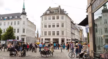 Et livlig europeisk gatebilde med folk som går og sykler på brostein. Bygningene rundt er klassiske og utsmykkede, med en fremtredende bygning med et spir. Kaféer og butikker ligger langs gaten, og flere trær gir grønt.