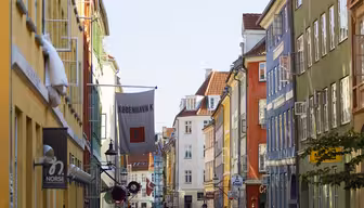 Et levende gatebilde i København med fargerike bygninger på hver side. På et skilt står det «København K» og folk går langs fortauet. Himmelen er klar og blå, og flagg henger over gaten.