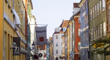Et levende gatebilde i København med fargerike bygninger på hver side. På et skilt står det «København K» og folk går langs fortauet. Himmelen er klar og blå, og flagg henger over gaten.