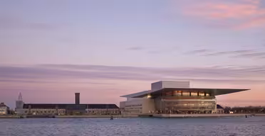 Copenhagen Opera House i skumringen med en rolig vannkant i forgrunnen. Himmelen er malt i pastellnyanser av rosa og lilla, og kaster en myk glød på den moderne arkitekturen og omkringliggende bygninger.