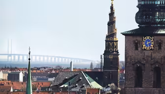 Utsikt over et bybilde med et fremtredende klokketårn med en blå og gullskive. I bakgrunnen er det en bro som strekker seg over vannet. Scenen inkluderer forskjellige hustak og et annet tårn med et spiralspir.