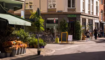 Et solrikt gatehjørne med en blomsterbutikk som viser potteplanter og buketter. Sykler er parkert utenfor en butikk som heter "DORA." Et tavleskilt har en tegning av en iskrem. Fotgjengere går forbi, og bygninger har grøntområder og markiser.