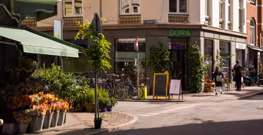 Et solrikt gatehjørne med en blomsterbutikk som viser potteplanter og buketter. Sykler er parkert utenfor en butikk som heter "DORA." Et tavleskilt har en tegning av en iskrem. Fotgjengere går forbi, og bygninger har grøntområder og markiser.