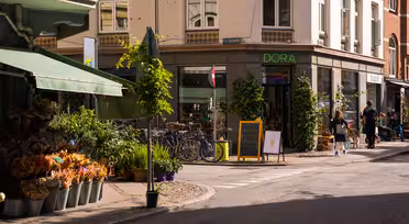 Et solrikt gatehjørne med en blomsterbutikk som viser potteplanter og buketter. Sykler er parkert utenfor en butikk som heter "DORA." Et tavleskilt har en tegning av en iskrem. Fotgjengere går forbi, og bygninger har grøntområder og markiser.
