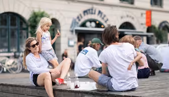 Folk sitter på en treplattform i urbane omgivelser. En kvinne med solbriller slapper av mens et barn i stripete antrekk står i nærheten. Sykler står i bakgrunnen foran en bygning med buet fasade.