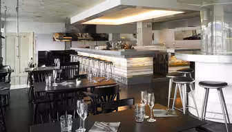 Moderne restaurantinteriør med svarte bord og stoler, en elegant bar med metallkrakker og synlig tak. Vinglass og servietter er dekket på bordene. Atmosfæren er minimalistisk og industriell.