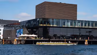 Moderne bygg med en mørk, strukturert fasade og store vinduer med utsikt over vannkanten. En båt passerer i forgrunnen på en solrik dag, med noen få mennesker som går langs kaien.