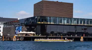 Moderne bygg med en mørk, strukturert fasade og store vinduer med utsikt over vannkanten. En båt passerer i forgrunnen på en solrik dag, med noen få mennesker som går langs kaien.