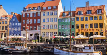 En livlig havnescene i København med fargerike bygninger i rødt, gult og grønt. Fortøyde båter langs kanalen under en klarblå himmel, med fortauskafeer og folk som nyter den sjarmerende atmosfæren.