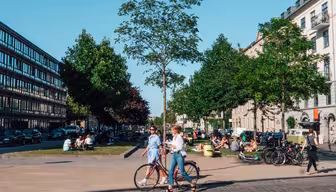 En yrende urban scene skildrer mennesker som går og sykler i en allé med solfylt vær. Grupper samles i et gressområde. Biler står parkert langs veien, og bygninger flankerer begge sider av gaten.