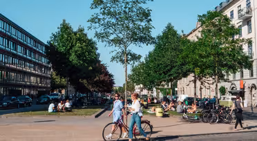 En yrende urban scene skildrer mennesker som går og sykler i en allé med solfylt vær. Grupper samles i et gressområde. Biler står parkert langs veien, og bygninger flankerer begge sider av gaten.
