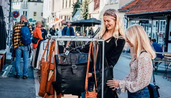 To kvinner ser på skinnvesker på et stativ ved gaten på et yrende bymarked. Folk er synlige i bakgrunnen og går langs den trange gaten med butikker og uteservering. Atmosfæren er livlig og uformell.