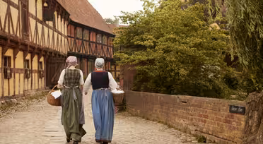 To kvinner i tidstypiske kostymer går på en brosteinsgate i en gammel landsby. De bærer vevde kurver og har lange skjørt, forklær og hodeskjerf. Gaten er omkranset av historiske tømmerhus og grøntområder.
