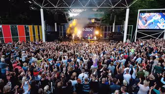 Et stort publikum samles utendørs foran en scene med levende lys og et band som opptrer. Folk rekker energisk opp hendene. Bakteppet viser ordet "THE BEAT" med fargerike paneler rundt scenen.