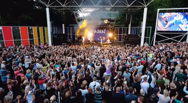 Et stort publikum samles utendørs foran en scene med levende lys og et band som opptrer. Folk rekker energisk opp hendene. Bakteppet viser ordet "THE BEAT" med fargerike paneler rundt scenen.