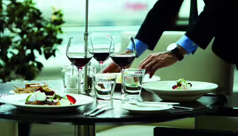 En person i dress dekker bord på en restaurant. På bordet står tallerkener med gourmetmat, fire glass rødvin og to glass vann. En liten plante er i bakgrunnen. Innstillingen fremstår elegant og eksklusiv.