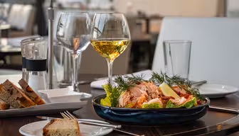 Et spisebord satt med et fat med salat toppet med laks, sitronskiver og dill. To glass hvitvin, en liten tallerken med brødskiver, og en pepperkvern står også på bordet. Atmosfæren er elegant og sofistikert.