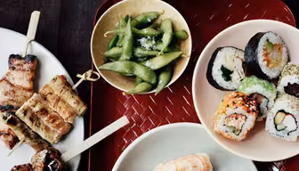Et arrangement av sushiruller, nigiri med reker og tunfisk, grillet kjøtt på spyd, edamame og en skål med ris på et mørkt bord. Røde spisepinner hviler på et levende vevd brett.
